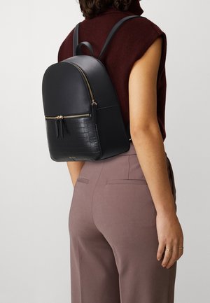 Femme portant un haut bordeaux sans manches et un pantalon mauve, avec un sac à dos en cuir noir doté de fermetures éclair dorées et d'une poche avant texturée façon crocodile.