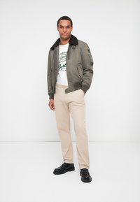 Olivengrønt bomberjakke med en brun pelskrave, hvid grafisk t-shirt under, og lysebeige bukser; sorte sko fuldender outfittet.