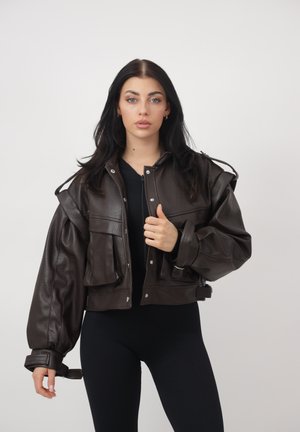 Vrouw met lang donker haar draagt een oversized donkerbruine leren jas over een zwart outfit, staand tegen een effen lichte achtergrond.