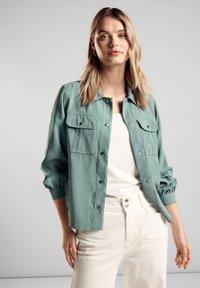 Světle zelená bunda na knoflíky s dvěma předními kapsami, dlouhými rukávy a elastickými manžetami, nošená přes bílou košili a světlé kalhoty.