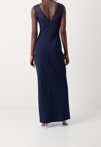 Marineblauwe avondjurk met een V-halsaccent van kant en een mouwloze ontwerp. Voorzien van een ritssluiting en een lange, aansluitende silhouette.