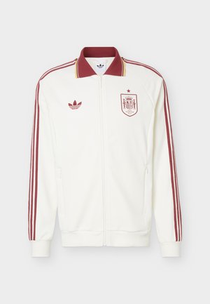 Chaqueta deportiva blanca con cremallera, con rayas color granate en las mangas, cuello granate, logotipo de Adidas en el pecho izquierdo y escudo de la selección nacional de fútbol de España en el pecho derecho.