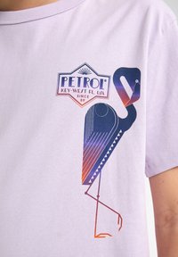 Lilla T-shirt med korte ærmer, der viser en grafik af en stiliseret fugl, ledsaget af mærkenavnet "PETROL" og lokationstekst.