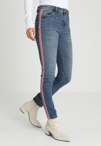 Blå denimjeans med röda och vita sidostriped, slim fit-design, hög midja, bärs med krämfärgade ankelstövlar.