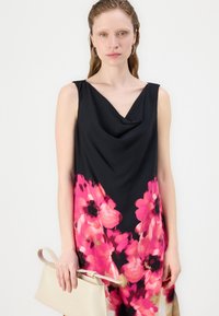 Rochie neagră fără mâneci cu un decolteu drapat în formă de cowl, având mari modele florale roz. Modelul ține o geantă bej.