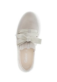 Beige mockasinsneaker med rund tå, prydd med en texturerad tygrosett framtill och en flexibel vit sula. Insidan visar varumärkeslogga.