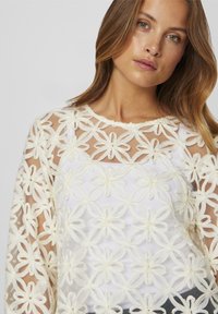 Haut transparent blanc avec broderies florales et pétales en 3D, sur une base blanche unie. Manches longues, texture légère.