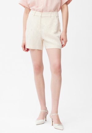 Vrouw draagt een lichtroze blouse, gestructureerde witte op maat gemaakte shorts en witte hoge hakken met puntige neus en enkelbandjes.