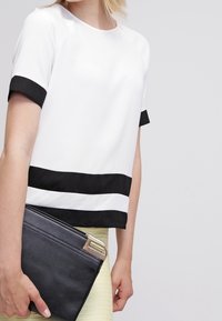 Haut blanc avec des accents noirs aux manches et à l'ourlet, associé à une pochette noire. Tissu lisse, coupe carrée et manches courtes.