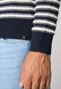 Prim-plan al unei mâini tatuate, purtând un pulover marin cu dungi albastre și albe și blugi de denim de culoare bleu deschis.