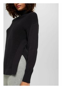 Maglione nero lavorato a maglia con collo alto, texture a coste, maniche lunghe e spacchi laterali. Abbinato a pantaloni grigi.