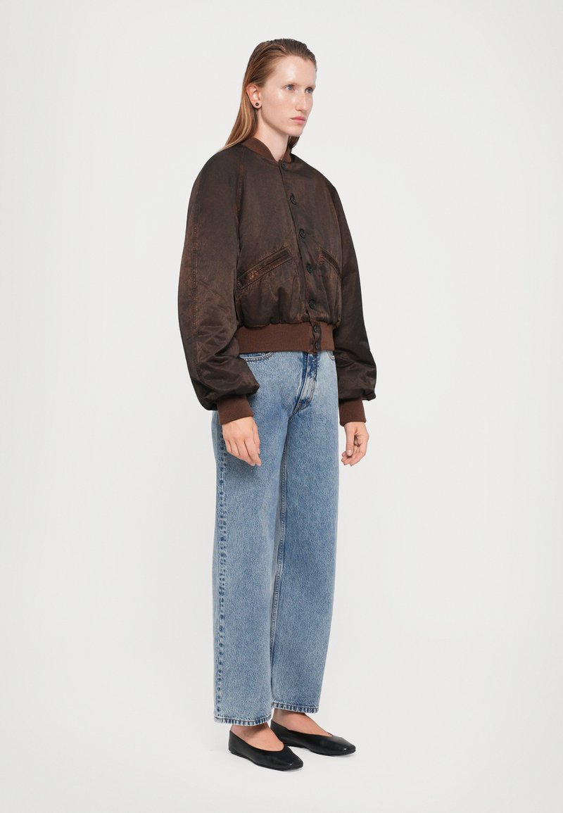 Veste bombardier marron avec de grands boutons et deux poches avant, associée à un jean large bleu clair et des ballerines noires.