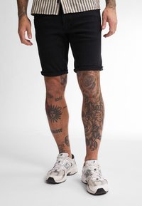 Shorts di jeans neri con risvolti, abbinati a sneakers bianche. Visibili diversi tatuaggi intricati sulle gambe, con fiori e disegni.