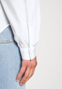 Chemise blanche à manches longues avec un poignet à bouton, texture lisse et coupe ajustée, associée à un jean en denim bleu clair. Bouton du poignet visible.