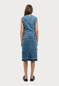Denim prsluk i suknja u srednje plavoj boji. Ima detalje s neurednim rubovima, bez rukava i okrugli žuti akcent na leđima.