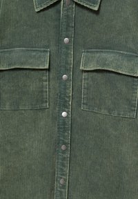 Chemise en velours côtelé vert avec des boutons-pression au centre et deux poches poitrine à rabat.