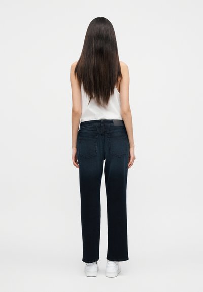 Femme aux longs cheveux foncés portant un haut blanc sans manches, un jean droit bleu foncé et des baskets blanches, debout de dos.