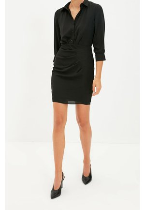 Kobieta ubrana w czarną sukienkę-koszulę zapinaną na guziki z marszczonym stanem i czarne wysokie szpilki z ostrym noskiem, stojąca na białym tle.