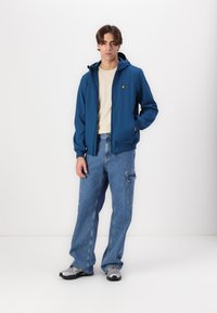Blå zip-up jacka med huva och gult logotyp, tillsammans med ljusblå denimbyxor med sidoficka och gråa träningsskor.