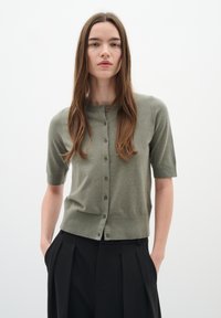 Cardigan kaki a maniche corte con colletto rotondo, dotato di chiusura a bottone e texture lavorata a maglia, abbinato a pantaloni neri larghi.