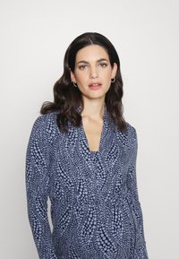 Vestido envolvente com padrão azul, featuring mangas longas e decote em V, feito de um tecido suave e com detalhe de cintura franzida.