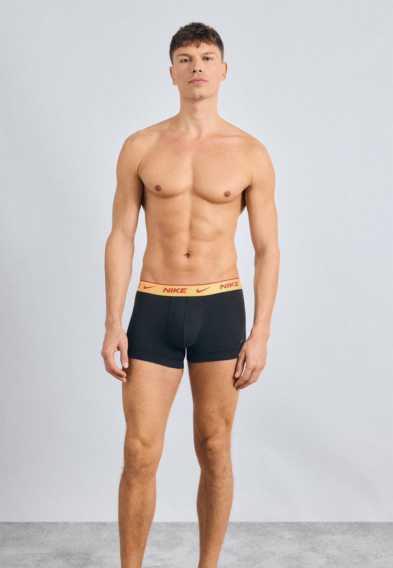 Čierne boxerky s elastickým pásom Nike v žltej a červenej farbe, s priliehavým strihom a hladkým materiálom, umiestnené na šedom pozadí.