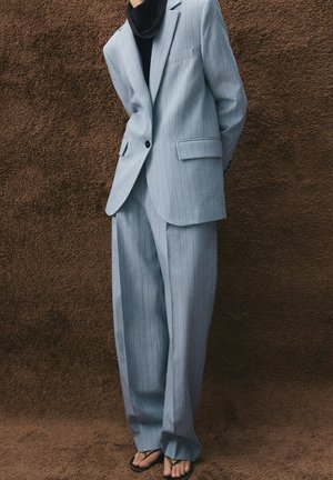Personne portant un costume à rayures fines bleu clair avec un blazer oversize et un pantalon large, associé à un haut noir et des sandales noires fines, debout sur un tapis marron.
