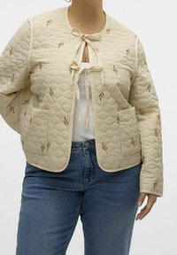 Gequilted crème jas met bruine bladborduursels, striksluiting bij de halslijn, ronde kraag en twee voorzakken, gecombineerd met blauwe jeans.