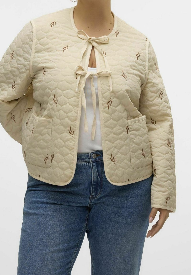 Gequilted crème jas met bruine bladborduursels, striksluiting bij de halslijn, ronde kraag en twee voorzakken, gecombineerd met blauwe jeans.