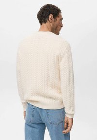 Maglione lavorato a maglia color crema con motivo a treccia, collo rotondo e polsini e orlo a coste. Indossato con jeans di denim azzurro chiaro.