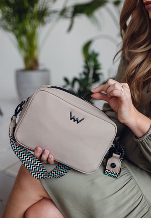 Beige rechteckige Tasche mit strukturiertem Stoff, schwarzem Reißverschluss und einem gewebten Träger mit Zickzackmuster in Türkis-, Schwarz- und Beigetönen.