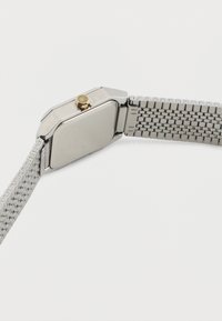 Τετράγωνο ασημένιο ρολόι χειρός με μεταλλικό μπρασελέ και χρυσό κουμπί, απεικονισμένο από την πίσω πλευρά σε λευκό φόντο.