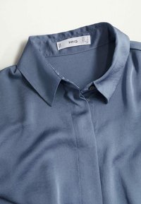 Chemise bleue à col avec boutons, étalée à plat, montrant la partie avant supérieure et l'étiquette de marque MNG avec les informations de taille à l'intérieur du col.