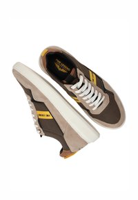 Bruine sneakers met witte veters, gemaakt van suède en canvas, met gele accenten en rubberen zolen. Heeft een logo en gestreept ontwerp aan de zijkanten.