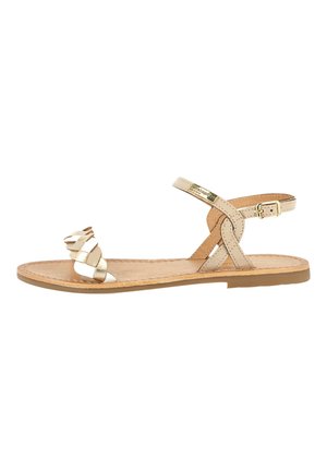 Flat sandal featuring a light beige leather base, braided gold straps, adjustable ankle buckle, and a textured sole.