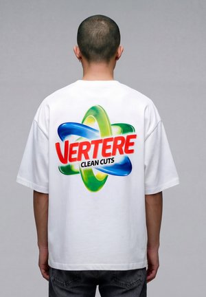 Person med kort hår iført en hvit T-skjorte med et fargerikt atomlignende design og ordene "VERTERE CLEAN CUTS" på baksiden.
