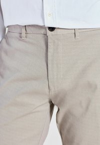 Pantalons à motif beige avec une surface texturée, dotés d'une fermeture à bouton et d'une coupe classique, partiellement visibles au-dessus d'une chemise blanche.