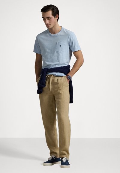 Light blue t-shirt with a chest pocket, beige linen trousers, navy sweater tied around waist, and navy sneakers. Simple, casual outfit.