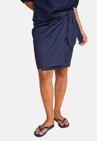 Marineblå omslagskjørt med knyttet detalj, laget av myk stoff. Kombinert med blå flip-flopper, som viser en avslappet sommerlook.