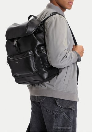 Uomo che indossa una giacca bomber grigio chiaro e jeans grigio scuro porta uno zaino grande in pelle nera con molte tasche con cerniera.