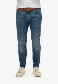 Vaqueros de denim azul con un corte ajustado, con ligero desgaste, combinados con un cinturón marrón y zapatillas en color crema.