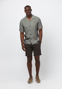 Korte mouwen grijs overhemd met knoopsluiting en een zak, gecombineerd met bruine opgerolde shorts. Model draagt bruine sneakers en staat op een effen achtergrond.