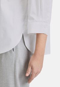 Weißes Langarmhemd mit Knopfmanschetten, mit abgerundetem Saum, kombiniert mit hellgrauen strukturierten Hosen. Aus weichem Baumwollmaterial.