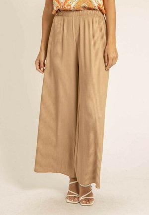 Femme portant un pantalon plissé ample beige, un haut à motif orange et des sandales blanches à brides sur un fond beige.