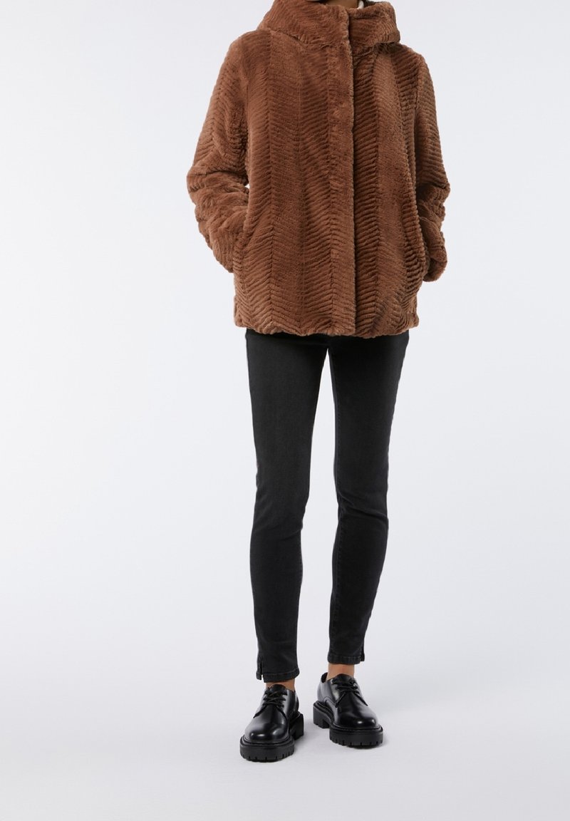 Giacca in pelliccia sintetica marrone con motivo a zigzag, colletto alto e chiusura con bottoni frontali, abbinata a jeans skinny neri e scarpe nere stringate.