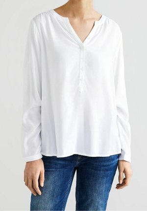 Kobieta ubrana w luźną, białą bluzkę z długim rękawem zapinaną na guziki z przodu, w połączeniu z niebieskimi dżinsami, stoi na jasnym tle.