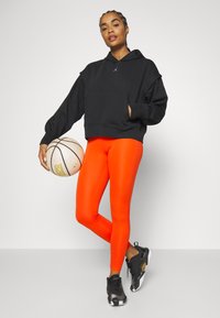 Felpa nera con cappuccio e tasca frontale, abbinata a leggings arancioni brillanti e scarpe da ginnastica nere con dettagli dorati. Tiene in mano un pallone da basket.