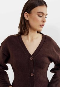 Dunkelbrauner Cardigan aus weichem Stoff, mit V-Ausschnitt, langen Ärmeln und zwei großen Knöpfen vorne. Strukturiertes Strickdetail an den Ärmeln.