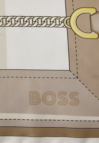 Material bej cu un design geometric, având o lanț de aur proeminent și accesorii metalice. Cuvântul "BOSS" este gravat într-o nuanță deschisă.
