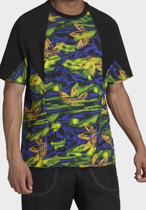 Homme portant un t-shirt noir à manches courtes avec un motif abstrait vert fluo, bleu et jaune et des logos adidas trefoil, associé à un pantalon noir.